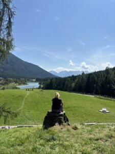 Frau sitzt auf Baumstamm und schaut in die Ferne. Dort sieht sie BErge